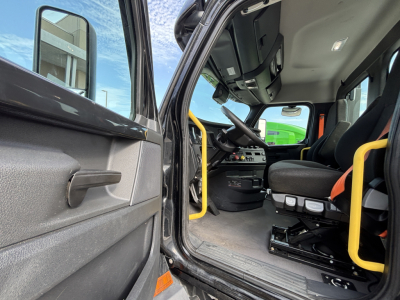 Cab interior of a Freightliner truck showing steering wheel, driver seat, yellow grab bars, dashboard, and open door with step.