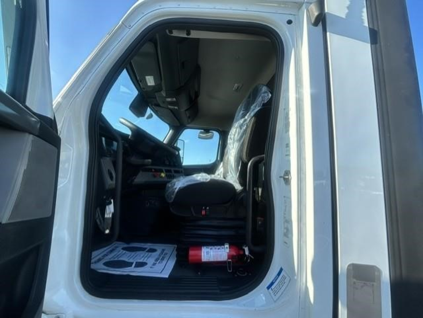 Cab interior of a Freightliner Cascadia 126 truck with open door, plastic-wrapped driver seat, steering wheel, dashboard, and a red fire extinguisher on the floor.
