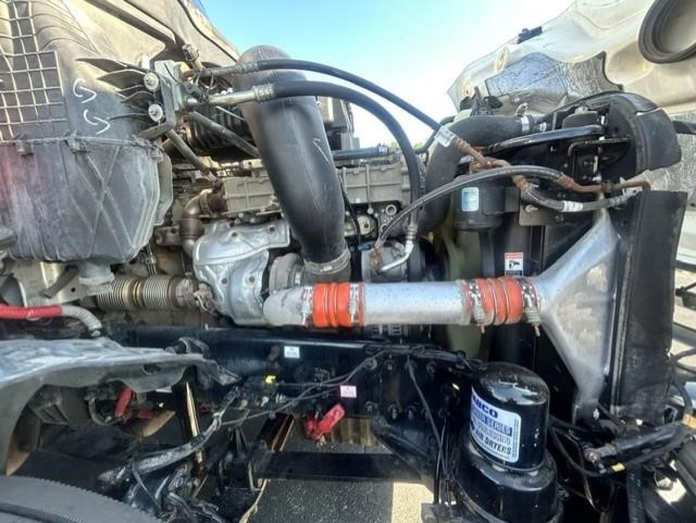 Engine bay of Freightliner Cascadia showing coolant hoses, filter, and various components. Visible labels include a blue and white filter canister and red/orange hose clamps.