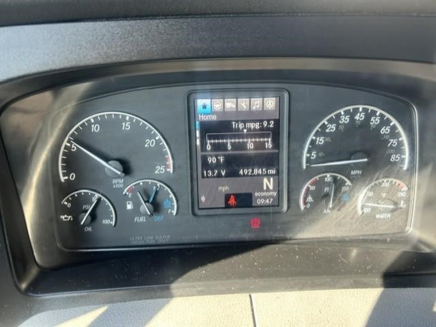 Dashboard of a Freightliner Cascadia showing instrument cluster with center digital display and gauges. Text on screen includes Home, Trip mpg: 9.2, 90°F, 13.7 V, 492.845 mi, ECONOMY 09:47. Left gauge shows mph and fuel; right gauge shows 