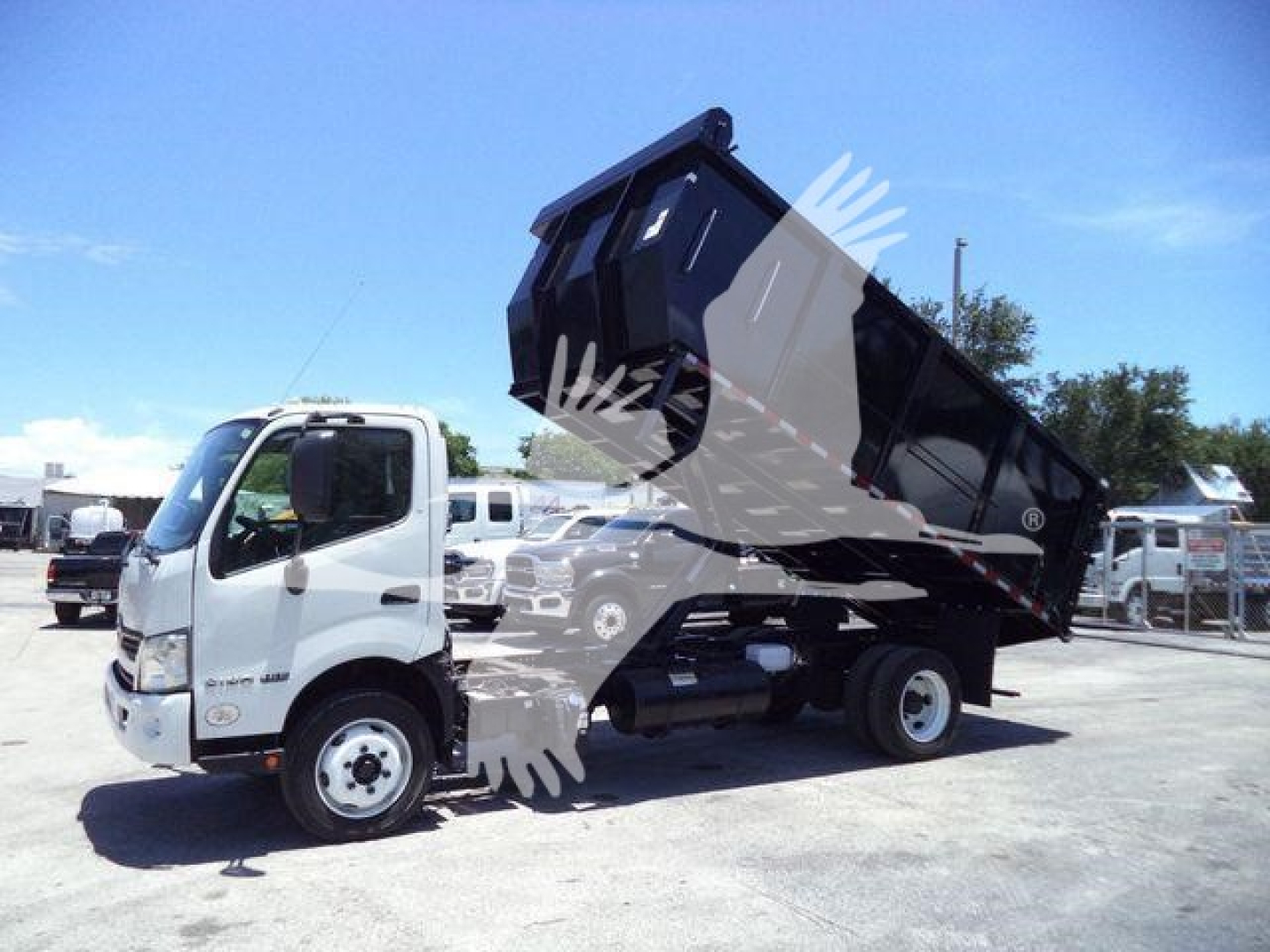 Truck dump bed raised showing Hino cab and dump body with hydraulic lift. Visible text: none legible; background shows other trucks.