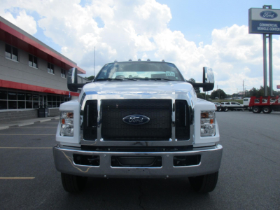 Front view of a white 2027 Ford F-750 SD cab and chassis truck in a dealership lot, large black grille with Ford emblem, chrome bumper, dual headlights, side mirrors.