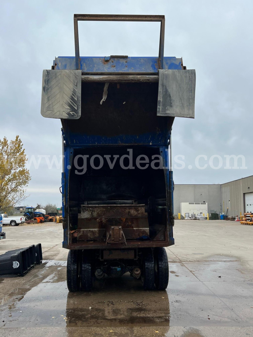 Rear view of a blue Peterbilt 320 dump truck with tailgate open, showing bed interior and undercarriage. Visible: two side steps, hydraulic components, and truck bed frame.