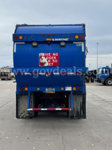 Rear view of a blue Peterbilt 320 dump truck showing Wayne body, taillights, mud flaps, and license plate area with stock number sticker.