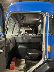 Peterbilt 320 cab interior viewed from open door showing driver's seat and dashboard with blue exterior cab, hanging overhead compartments, and a red toolbox on the floor.