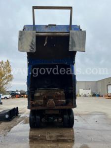 Rear view of a blue Peterbilt 320 dump truck with tailgate open, showing bed interior and undercarriage. Visible: two side steps, hydraulic components, and truck bed frame.