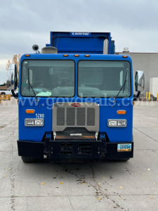 Front view of a blue Peterbilt 320 garbage truck showing grille, headlights, bumper, and cab ID; visible vehicle is in-service.