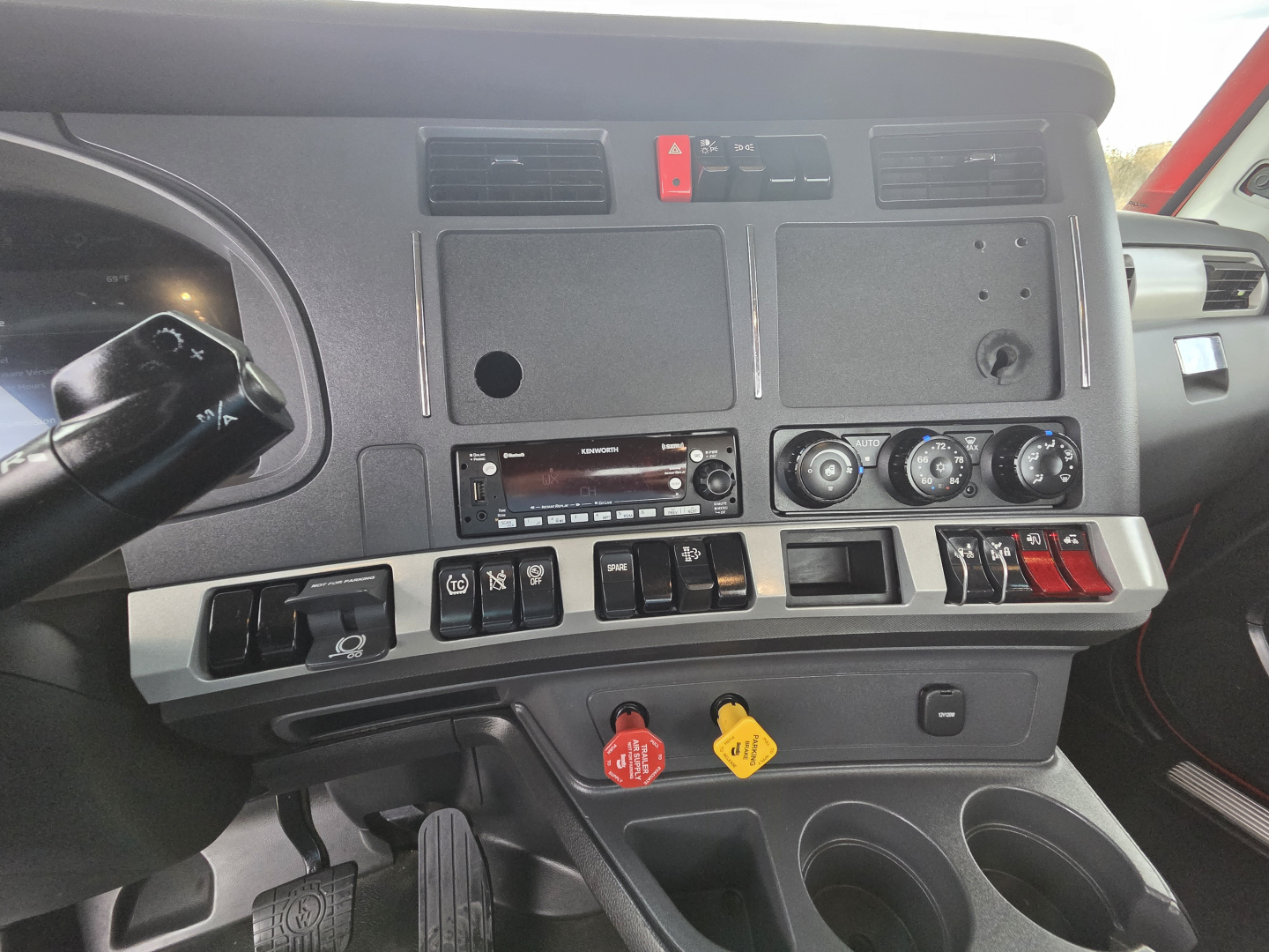 Truck cab dashboard interior with multiple switches, gauges, and vents visible; center console area shows climate controls and warning lights; red hazard button and yellow/red knobs.