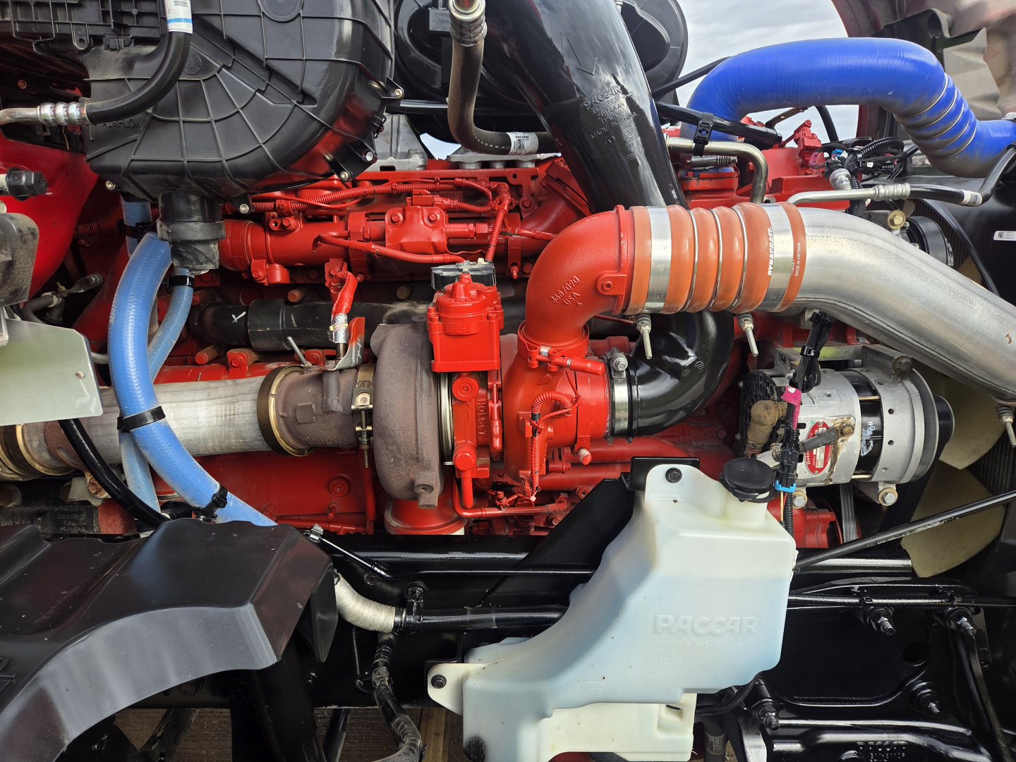 Kenworth T680 engine bay showing red engine components, blue hoses, and a large silver exhaustpipe with orange heat shield.