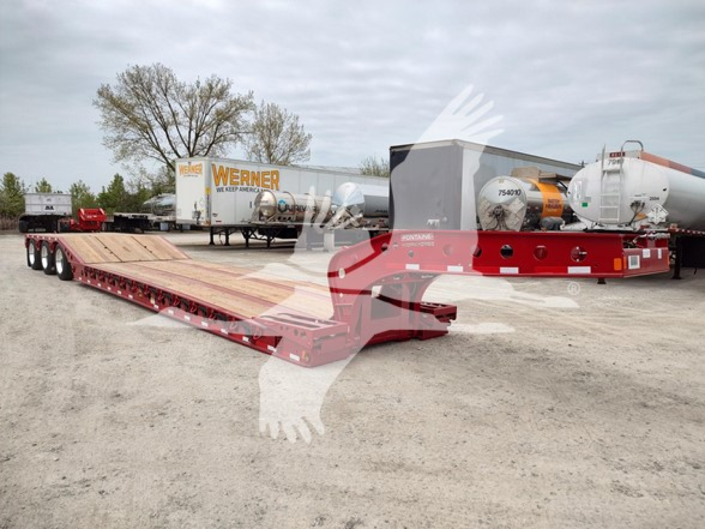Fontaine paver lowboy trailer with red kingpin area and long flatbed; background shows semi-trailers and industrial equipment.