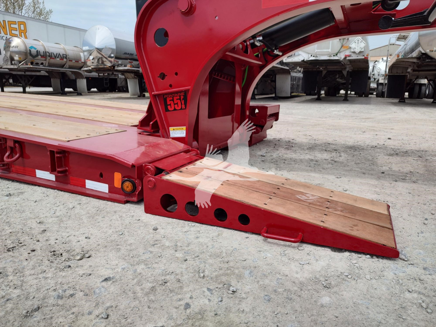 Fontaine paver pro lowboy trailer showing red steel frame and wooden loading ramps with holes and cutouts, side labels, and surrounding equipment in lot.