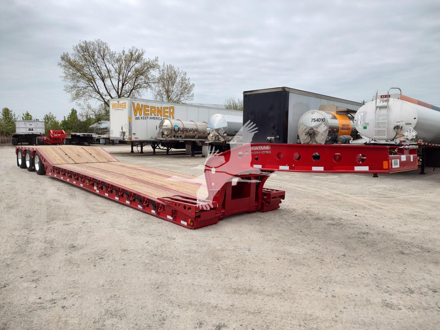 Lowboy trailer deck with red framing and long wooden surface, Fontaine Paver Pro trailer in a yard with other trailers and tanks in background.