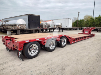 Fontaine paver lowboy trailer with red frame and long platform, three axles, chrome wheels, and wooden deck; appears new with stock display in yard