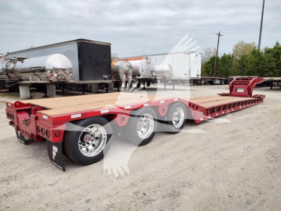 Fontaine paver lowboy trailer with long red frame and multiple axles, wooden deck, parked in yard with other trailers in background.