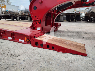 Fontaine paver pro lowboy trailer showing red steel frame and wooden loading ramps with holes and cutouts, side labels, and surrounding equipment in lot.