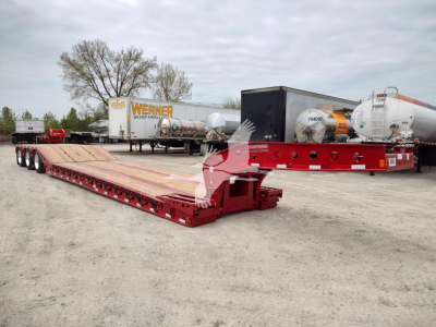 Lowboy trailer deck with red framing and long wooden surface, Fontaine Paver Pro trailer in a yard with other trailers and tanks in background.