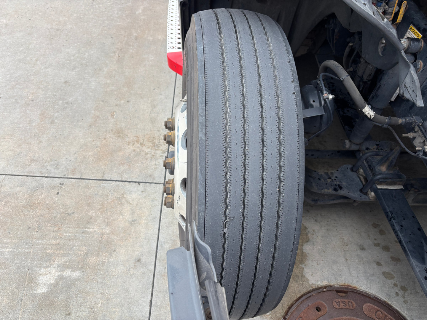 Truck wheel and suspension detail showing a single front drive tire with tread worn to a smooth center, rim with lug nuts, part of cab and chassis visible.