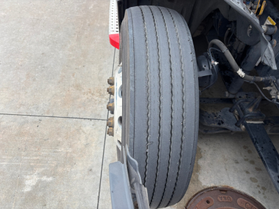 Truck wheel and suspension detail showing a single front drive tire with tread worn to a smooth center, rim with lug nuts, part of cab and chassis visible.