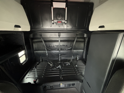 Truck cab interior showing sleeper area and cargo floor with straps and bulkhead behind. Visible branding on back wall and overhead storage panels.