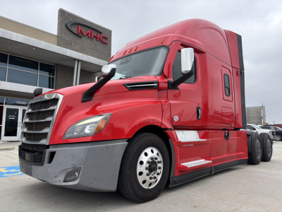 Red Freightliner PT126064ST semi-truck cab with large front grille and sleeper, dealership building in background, windowed cab door, chrome mirrors, white wheel rims.