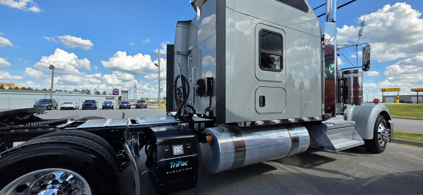 Truck cab and chassis area of a Kenworth W900 showing side fuel tanks, steps, and exhaust stack; no legible model text visible in this view.