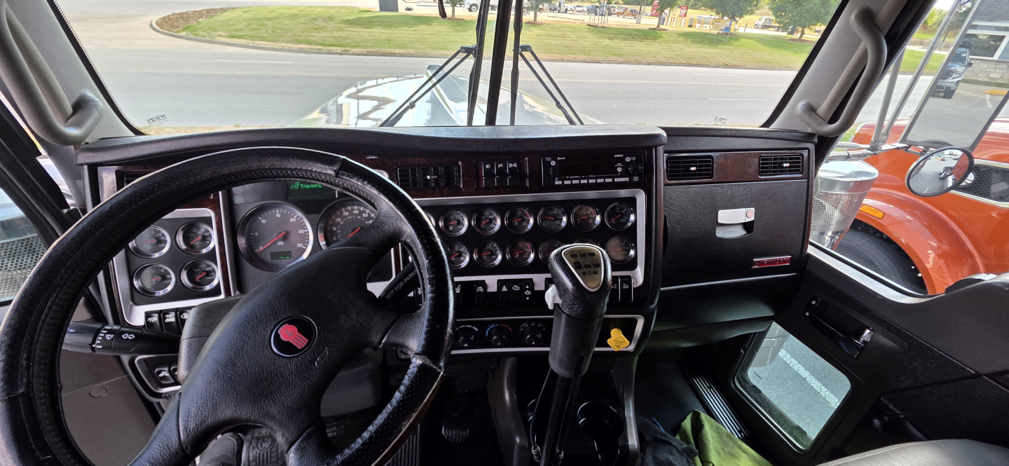 Kenworth W900 cab interior showing steering wheel, dash gauges, gear selector, and control panels.