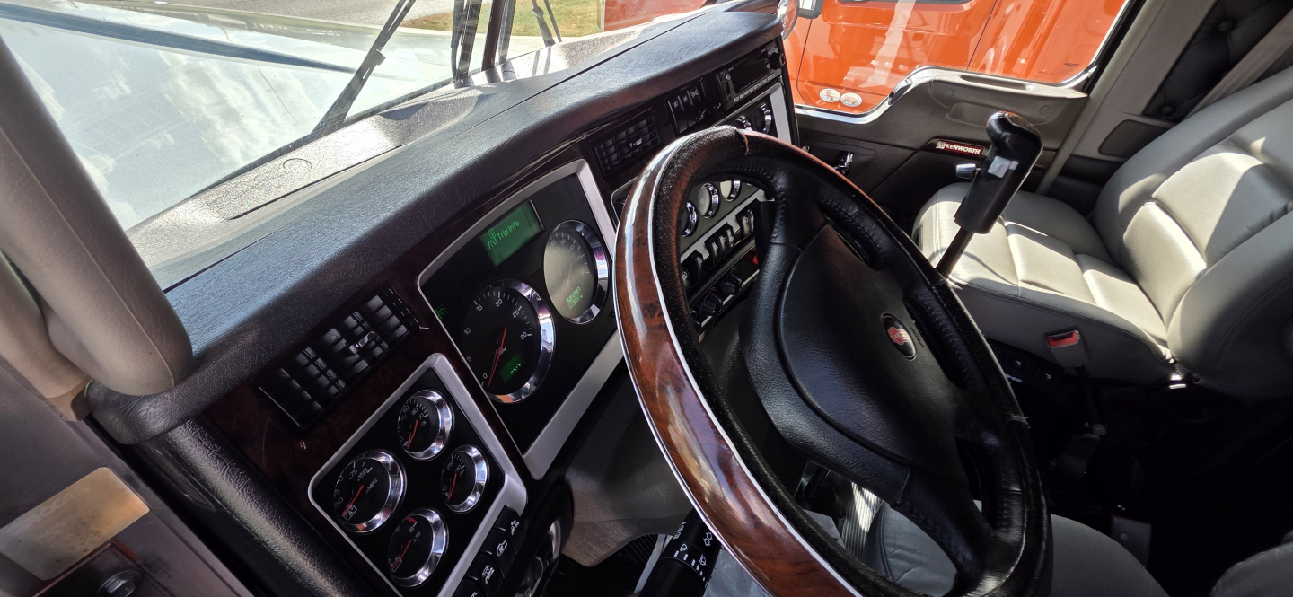 Cab interior of a Kenworth W900 showing steering wheel and dashboard with gauges and controls.