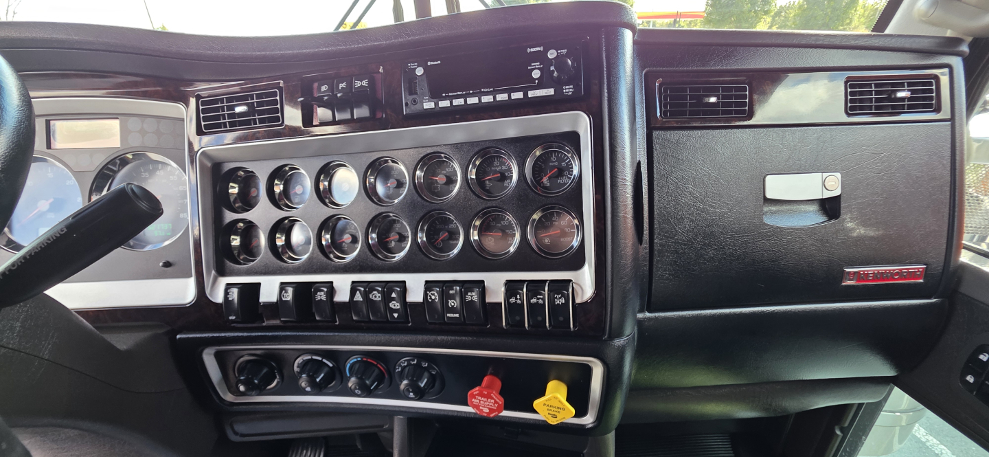 Kenworth W900 cab interior dashboard with multiple gauges and switches, steering wheel on left, center console with chrome dials and controls.