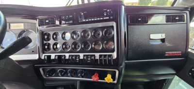 Kenworth W900 cab interior dashboard with multiple gauges and switches, steering wheel on left, center console with chrome dials and controls.
