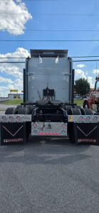 Kenworth W900 rear view showing chassis, dual rear axles, taillights, mud flaps with Kenworth branding, and a metallic cab-back with visible rear bumper and wheel wells.