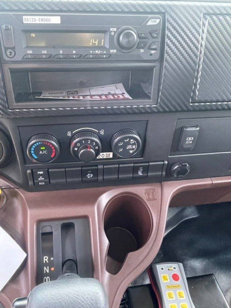 Dashboard interior of a 2027 Hino L7 showing radio unit, climate controls, and cup holder area; gear selector visible on left and brown center console.