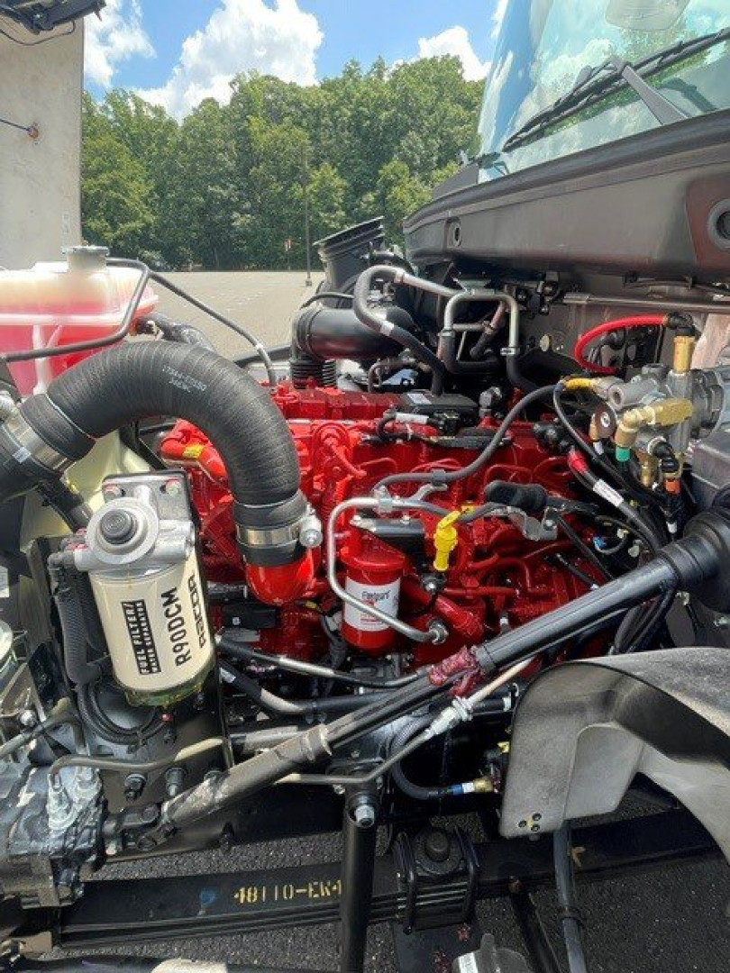 Engine bay of a 2027 Hino L7 hooklift truck showing red engine block, hoses, filters, and various metal lines with surrounding components.