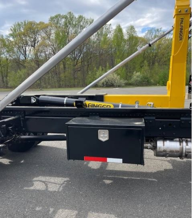 Hino L7 hooklift truck side view showing a black tool/storage box mounted under frame and hydraulic cylinders.