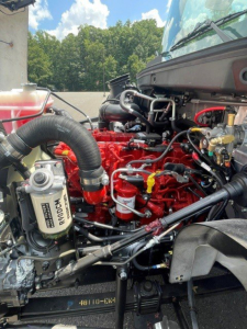 Engine bay of a 2027 Hino L7 hooklift truck showing red engine block, hoses, filters, and various metal lines with surrounding components.