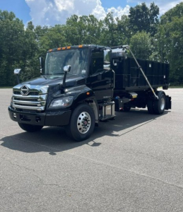 Truck cab and dump body of a 2027 Hino L7 hooklift truck, black color, front grille with Hino emblem, chrome bumper, side mirrors, and tag lines.