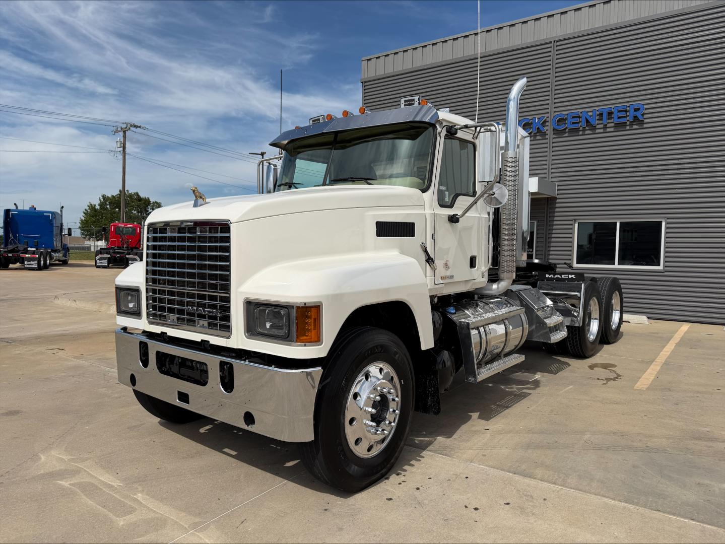 White 2019 Mack PI64T conventional daycab truck in a parking lot, with large chrome bumper, dual rear wheels, and Mack trucks center building in background.
