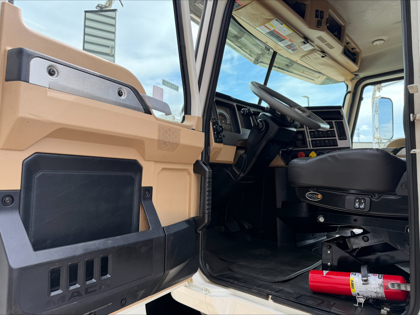 Cab interior of a 2019 Mack PI64T with open driver door showing steering wheel, dashboard, and seat; tan and black interior, red fire extinguisher mounted near the floor.