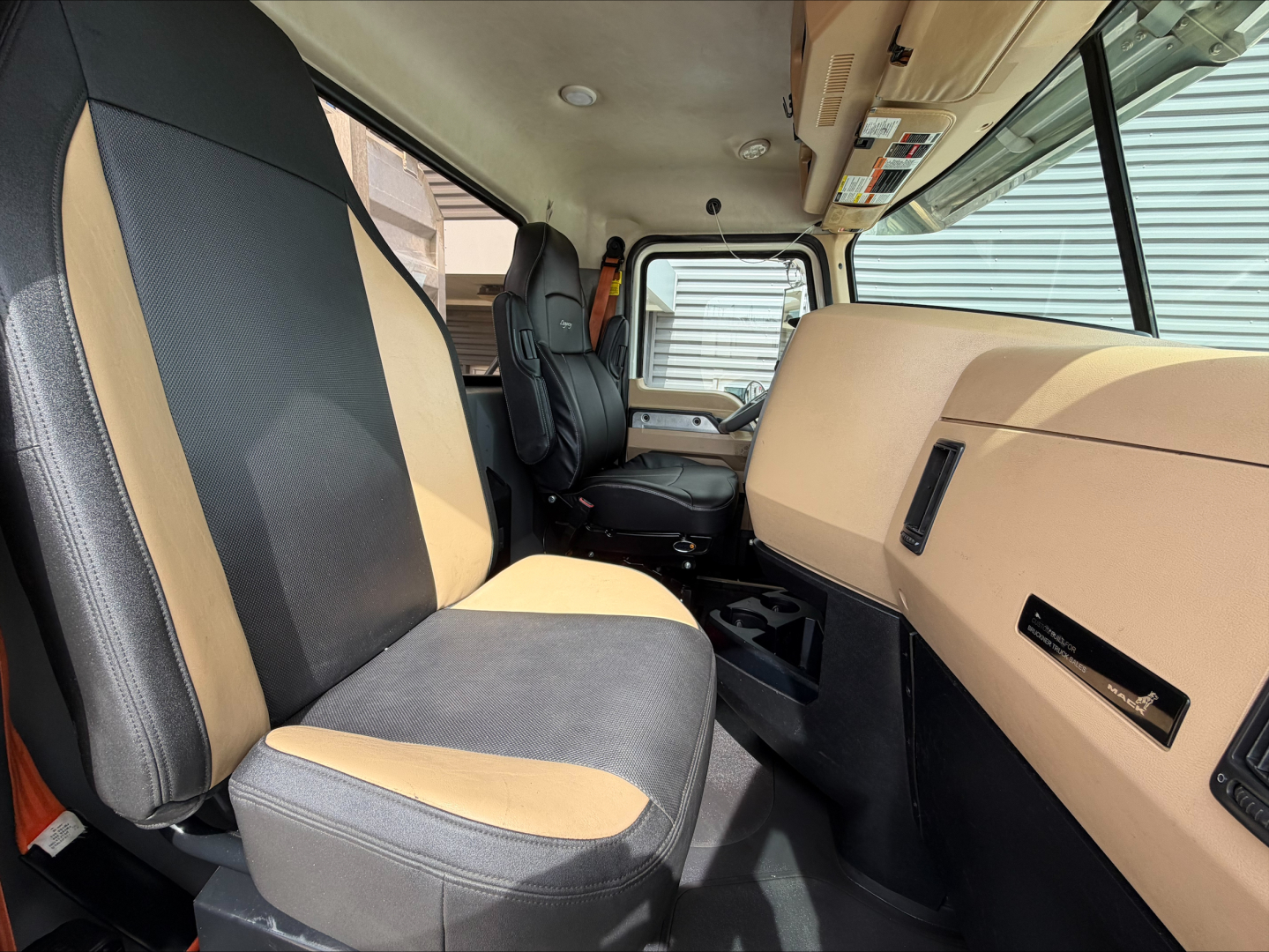 Truck cab interior of a 2019 Mack PI64T, front seats with two-tone upholstery, dashboard, and steering area visible.
