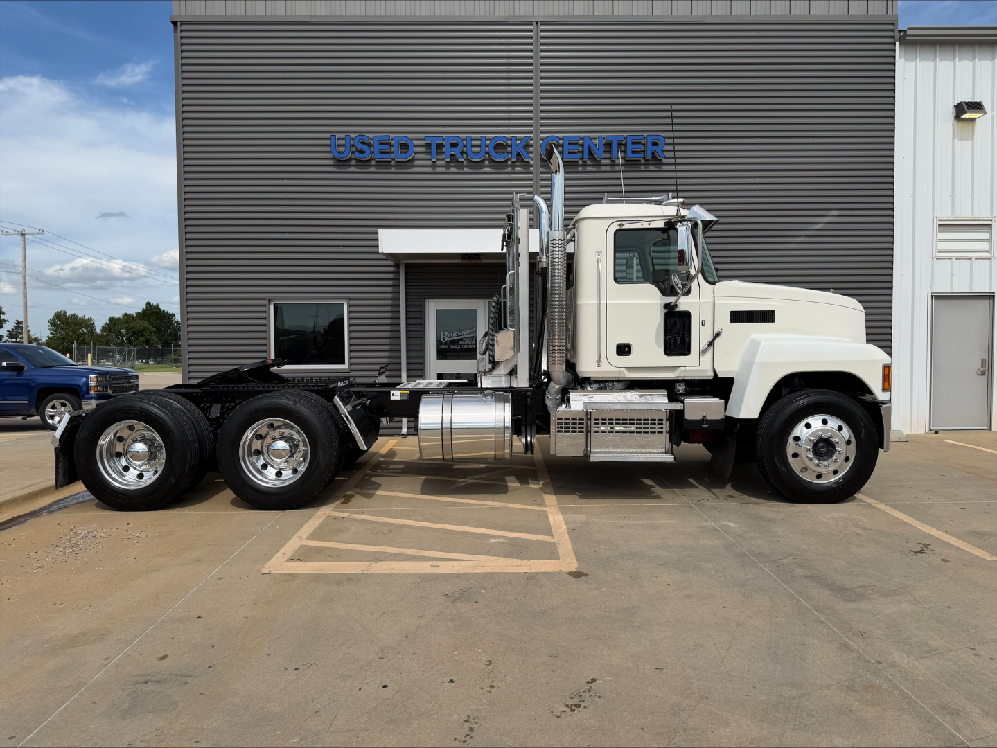 2019 Mack PI64T conventional daycab truck in side view. White cab, dual rear axles with chrome fuel tanks, stacked exhaust, parked in front of a building labeled 'USED TRUCK CENTER'