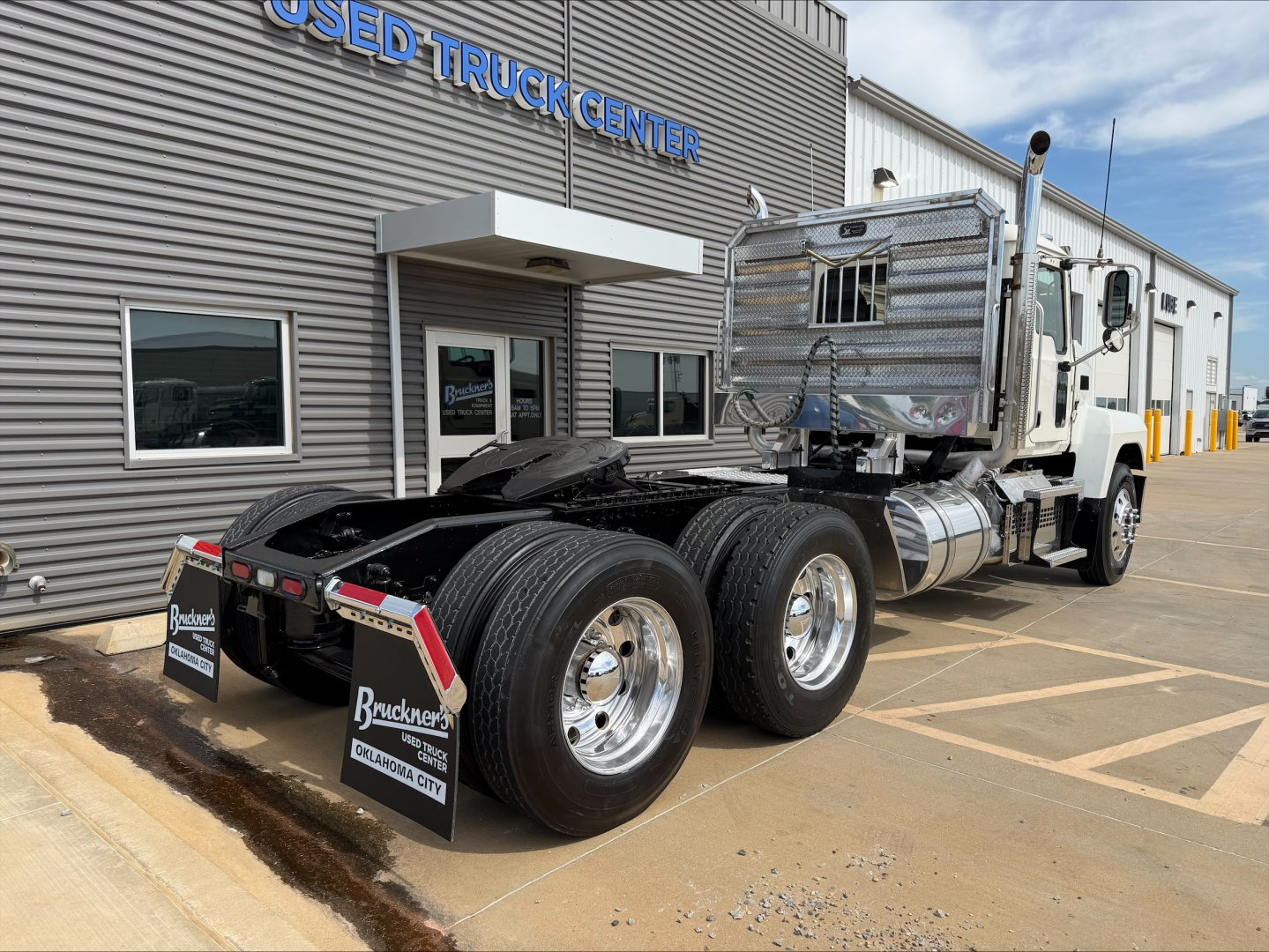 2019 Mack PI64T truck tractor in front of used truck center showing tandem rear axles and chrome fuel tanks.