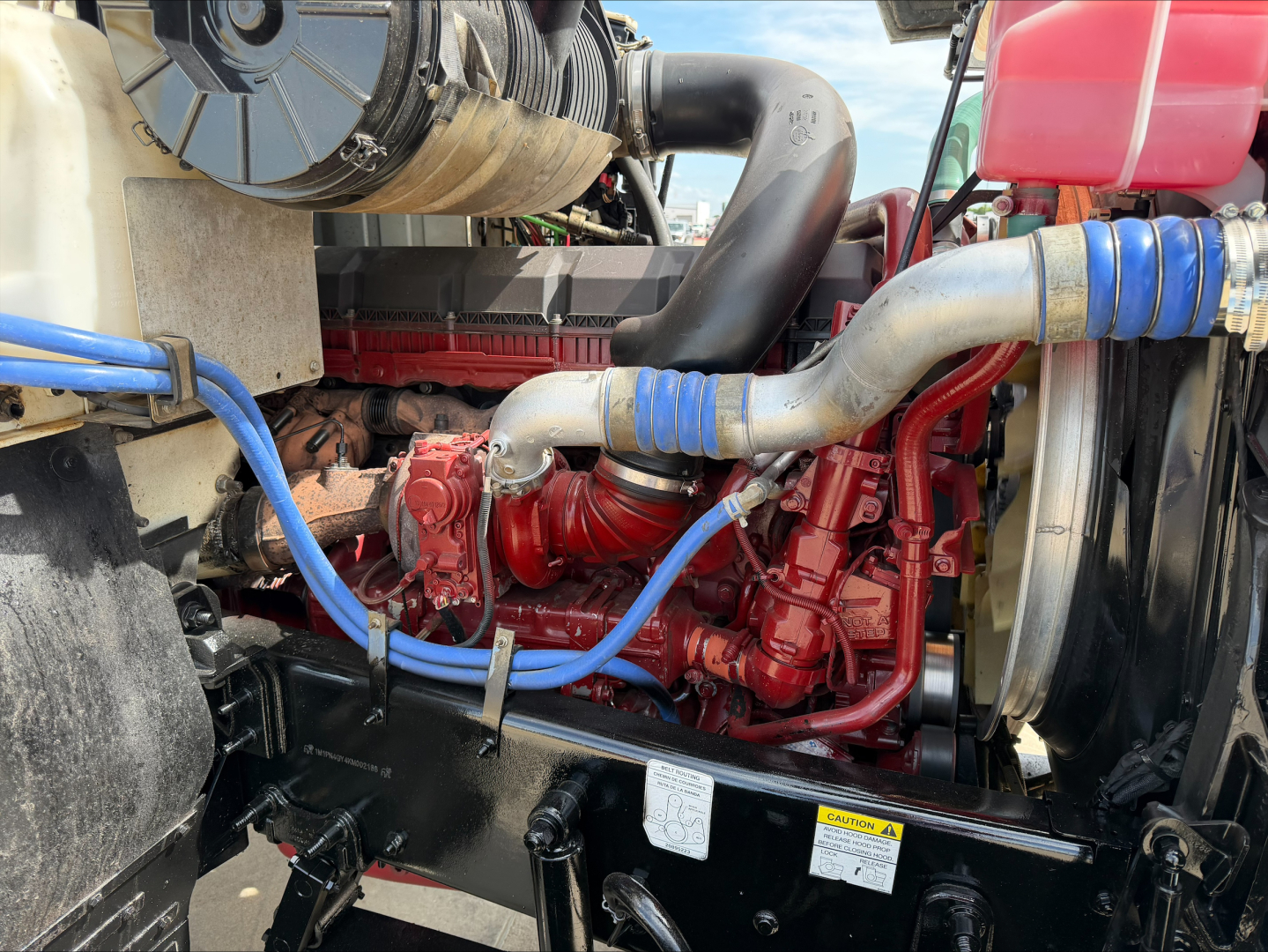 Mack PI64T engine bay showing red engine block with blue hoses, red coolant tank, air intake tubing, and surrounding black framework.