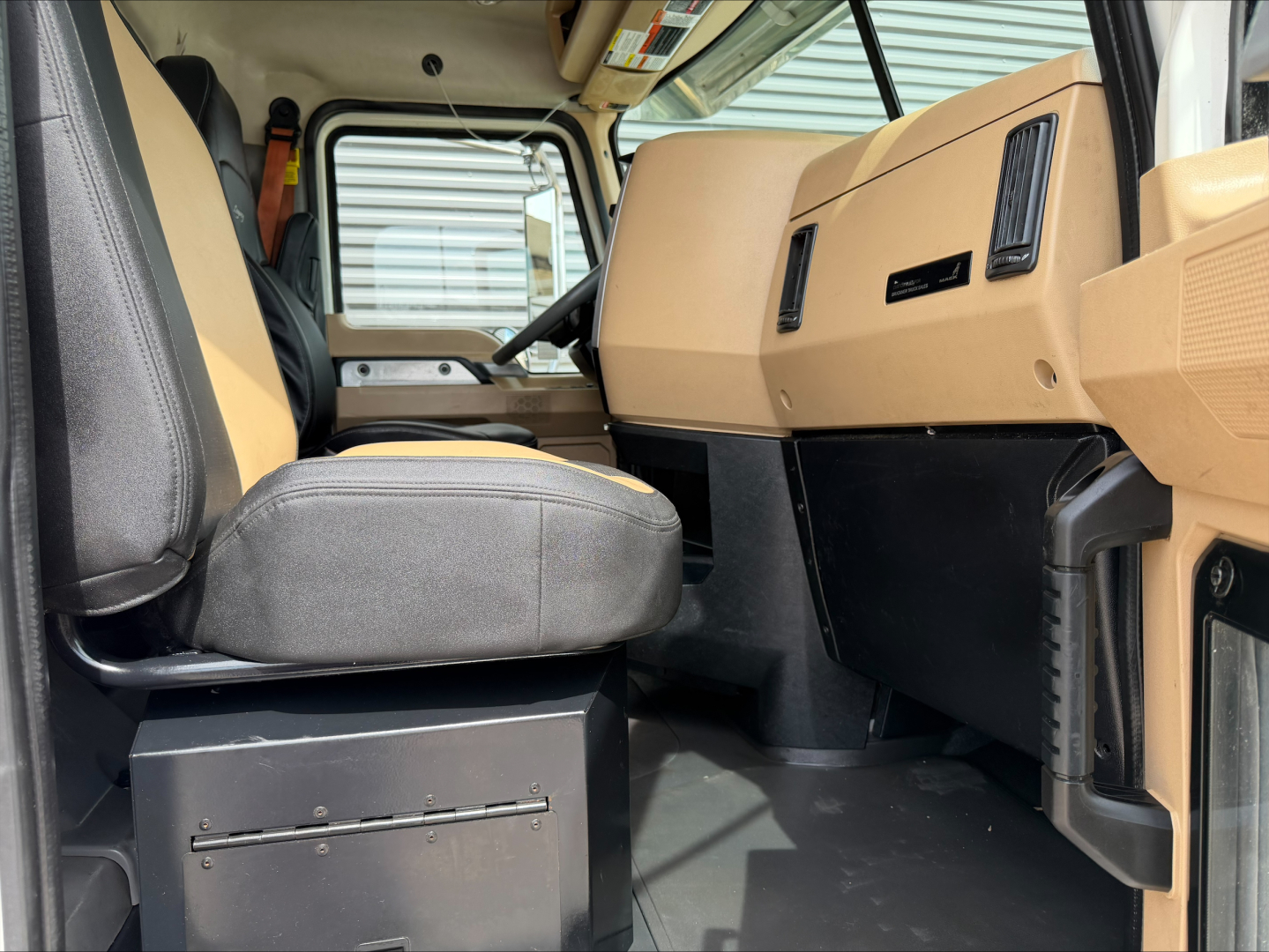 Interior of a 2019 Mack PI64T daycab showing brown dashboard, driver's seat, and steering wheel; front storage drawer under seat; vinyl floors; visible window and cabin controls.