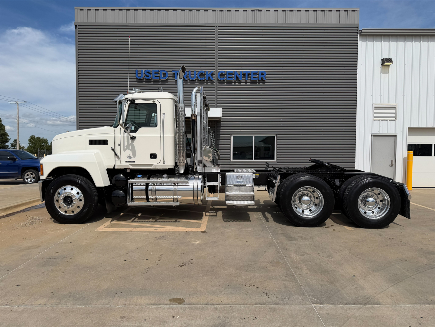 2019 Mack PI64T conventional daycab truck side view showing cab, chassis, fuel tanks, exhaust stacks, and rear tandem wheels in front of aUsed Truck Center building.