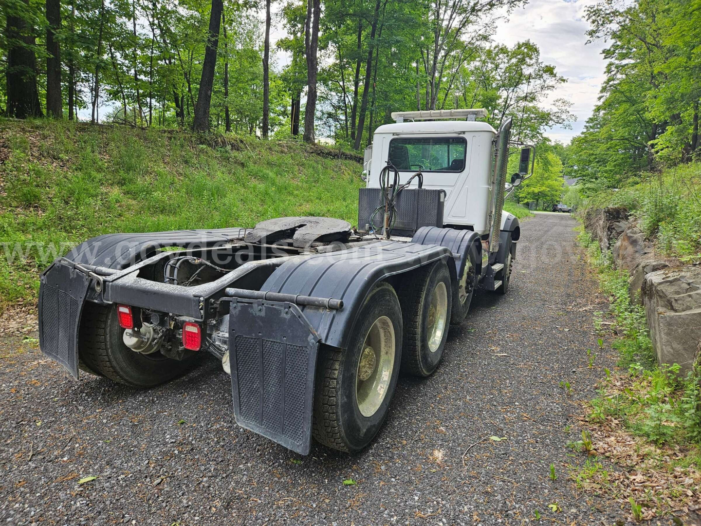 Mack GU813 tractor rear view showing dual rear axles on a gravel road in a wooded area; chassis and fifth wheel area visible with mudflaps.