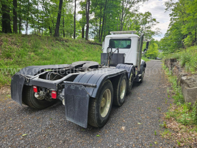 Mack GU813 tractor rear view showing dual rear axles on a gravel road in a wooded area; chassis and fifth wheel area visible with mudflaps.