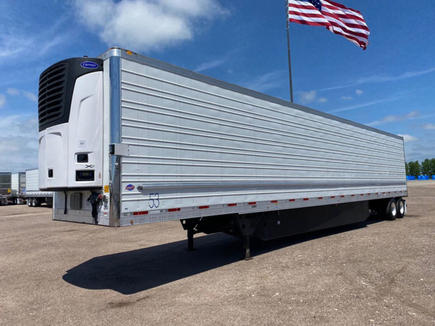 Refrigerated trailer with front-mounted refrigeration unit and silver corrugated side panels, parked on gravel with American flag in background.