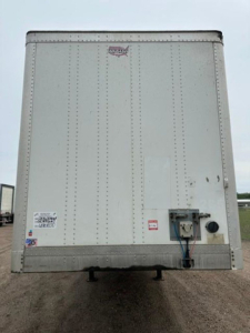 Rear view of a white Wabash van trailer with hitch and access panel; small decals near lower left corner and a black circular component with blue/black hoses on the right; no door open.