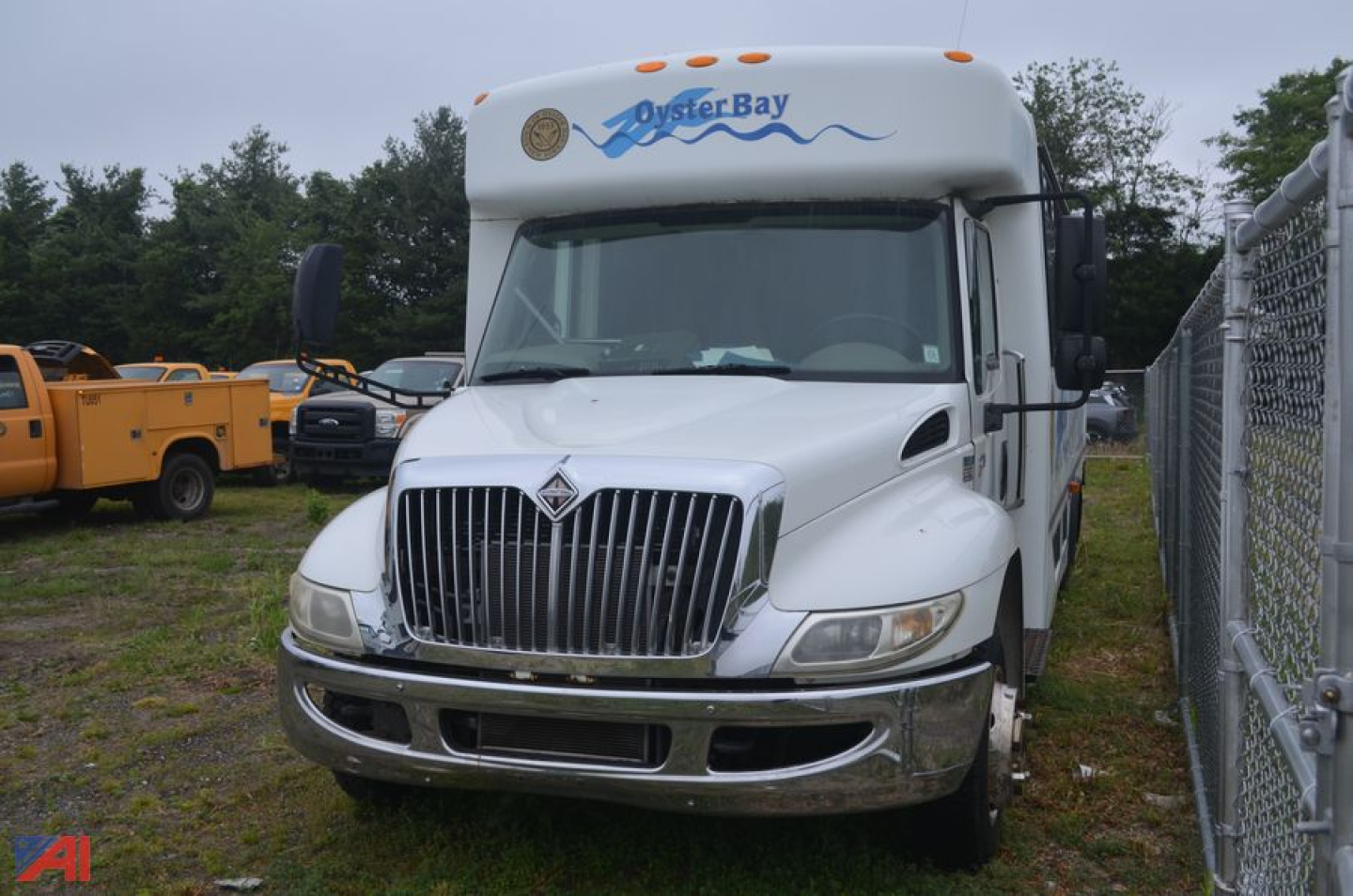 White 2009 International 3200 bus front view showing grille, headlights, and Oyster Bay graphic on cab, with chain-link fence on right.