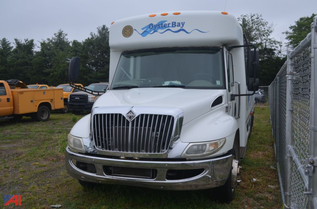 White 2009 International 3200 bus front view showing grille, headlights, and Oyster Bay graphic on cab, with chain-link fence on right.
