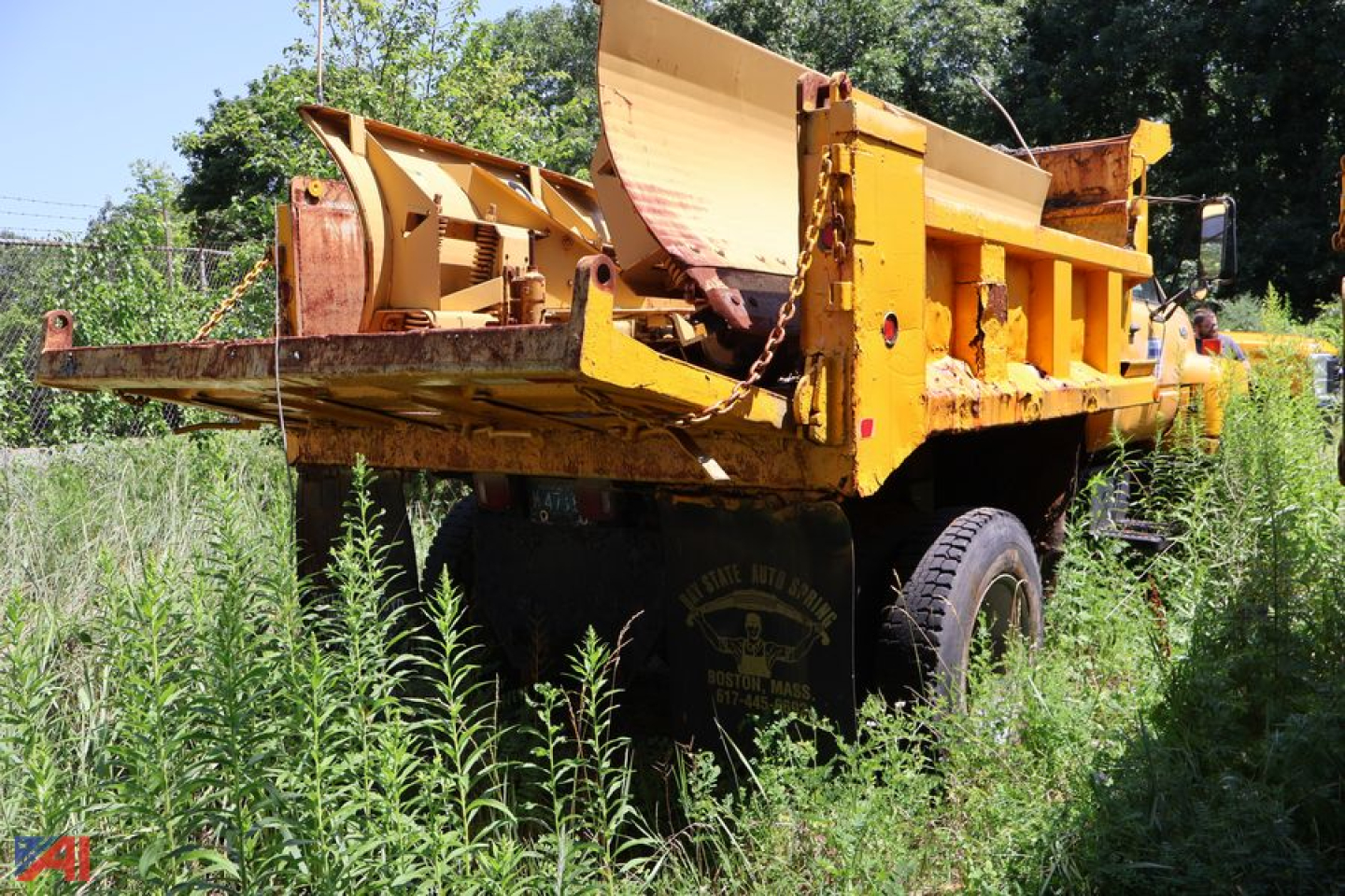Dump truck bed showing yellow Chevrolet Kodiak C7500 with overgrown grass around rear tires and chains on the bed.